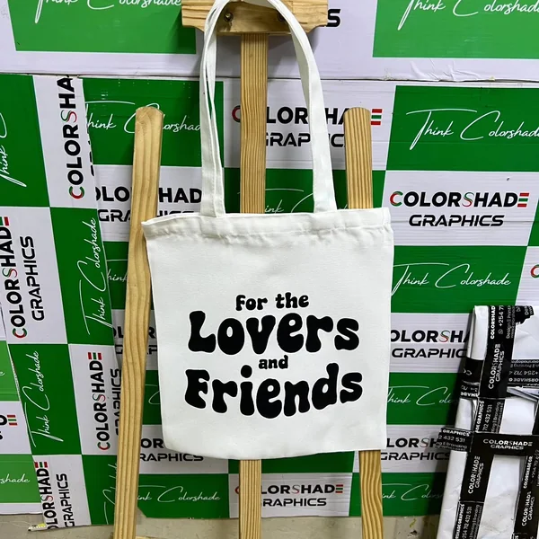 White canvas tote bag printed "For the Lovers and Friends", hung against a Colorshade backdrop